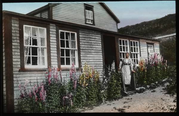 Woman by frame house; snapdragons in garden