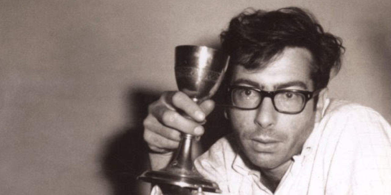 Black and white photograph of poet and writer Al Pittman. He is wearing glasses and a light-colored shirt, with slightly unkempt hair. He holds up a metal goblet in his right hand, gazing directly into the camera with an intense and contemplative expression. The background is plain and slightly blurred.