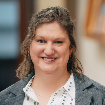 Portrait of Alison Carr smiling, wearing a light gray blazer over a white blouse with stripes, photographed indoors with a softly blurred background.