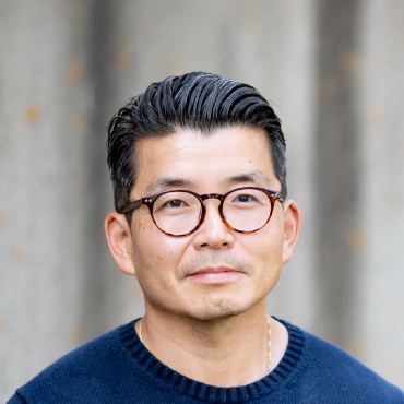 Portrait photo of Andrew Kim wearing glasses and a dark sweater, standing in front of a blurred background
