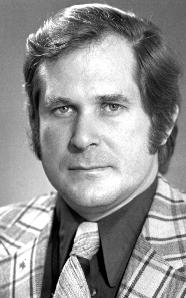 A black-and-white portrait of a middle-aged Angus Bruneau with light skin, short wavy hair, and sideburns. He has a confident, serious expression and is looking directly at the camera. He is wearing a plaid blazer, a dark collared shirt, and a textured tie. The background is plain and slightly blurred.