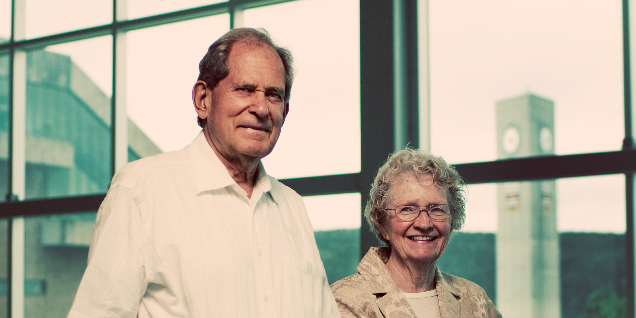 Angus and Jean Bruneau stand side by side, smiling, in a modern glass walkway with metal railings. Large windows behind them reveal a view of the Memorial University campus, including the iconic clock tower in the background. The man is wearing a short-sleeved white button-up shirt and dark pants, while the woman is dressed in a beige blazer over a light-colored top. They appear relaxed and conten