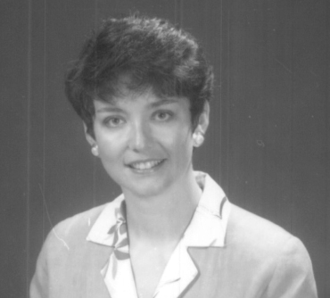 A black and white portrait of Victoria Collins with short, dark hair, smiling gently at the camera. She is wearing a light-colored blazer over a collared blouse. The background is plain and softly lit, giving the image a classic and professional appearance.