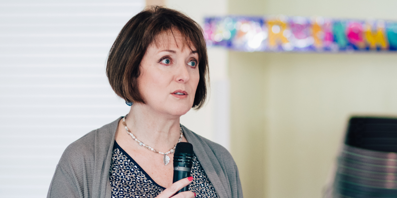 Victoria Collins, a woman with short brown hair, speaks into a handheld microphone while looking slightly to the side. She wears a patterned blouse, a gray cardigan, and a silver necklace with a pendant. Her expression is serious and focused. The background is slightly blurred, featuring a colorful banner and stacked chairs in a well-lit indoor space.