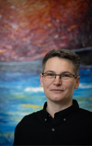 Ailsa Craig stands in front of a vibrant oil painting with red, blue, and yellow tones. They are wearing glasses and a dark button-up shirt, smiling softly at the camera.