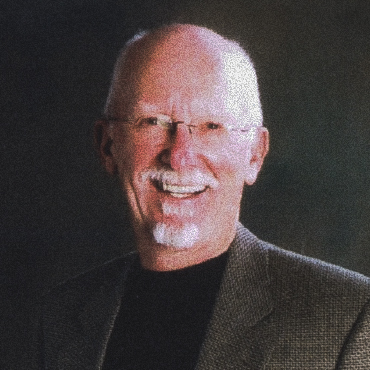 Portrait of Darryl Fry smiling, wearing glasses, a dark shirt, and a textured gray blazer, set against a dark studio background.