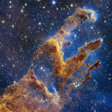 A stunning image of the Pillars of Creation, a formation of interstellar gas and dust located in the Eagle Nebula. The towering, finger-like structures glow with shades of gold and brown, set against a deep blue, star-filled cosmic backdrop. The edges of the pillars appear illuminated by radiation from nearby stars, giving them an ethereal, almost sculpted appearance.