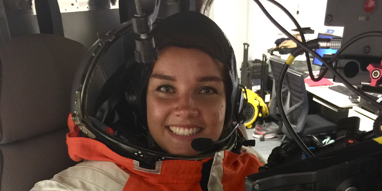 Bethany Downer, a young woman wearing a spacesuit, sits in a cockpit, smiling at the camera. She is wearing an orange and white suit with a patch that reads 