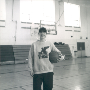 A black-and-white photo of Michelle Healey, a young undergraduate woman standing in a gymnasium, holding a basketball under one arm. She is wearing a sweatshirt with a Sea-Hawks logo and dark jogging pants. The background features basketball hoops, large windows, and empty bleachers, creating a classic sports setting. She has a friendly smile and a relaxed posture.