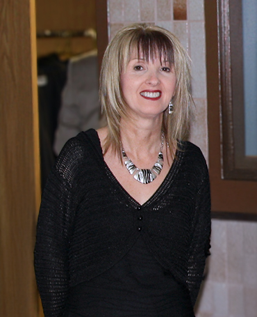 Deborah Janes, with shoulder-length blonde hair and bangs, smiles at the camera. She is wearing a black V-neck top with a subtle shimmer, a silver statement necklace, and matching earrings. The background features a tiled wall and a partially open wooden door with a coat hanging inside. The lighting is warm and soft.