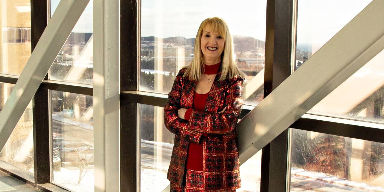 Deborah Janes, a woman with long blonde hair, stands with her arms crossed, smiling confidently at the camera. She is wearing a red and black plaid blazer over a red dress. The background features large windows with metal beams, showing a snowy landscape and a cityscape with hills in the distance. Natural light streams through the windows, casting a warm glow on her face.