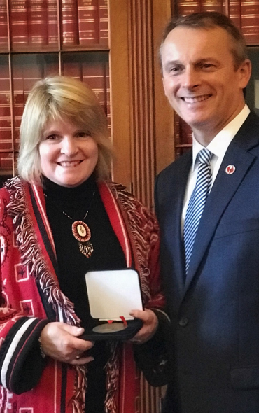 Brenda Jeddore, wearing in a red and black patterned jacket, holds an open award box, standing next to Senator David Wells dressed in a dark suit and tie. They are smiling, posing in front of a wooden bookshelf filled with red-bound books.