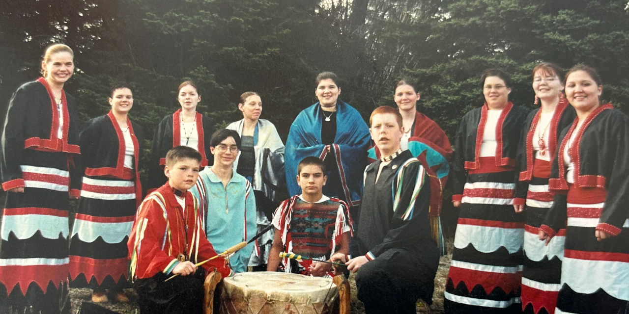 A group of Indigenous teenagers posing outdoors in traditional regalia. The group is dressed in colorful clothing featuring intricate patterns, fringes, and beadwork. Some individuals wear black, red, and white ribbon skirts and jackets, while others have shawls draped over their shoulders. Four boys sit at the front holding drumsticks, with a large floor drum between them. The background consists