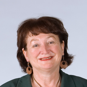 Color portrait of Joan Oldford with short brown hair, wearing earrings and a dark green top, smiling at the camera.