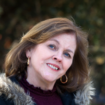 Color portrait of Lynn Morrissey with shoulder-length light brown hair, wearing hoop earrings, a burgundy top, and a fur-trimmed coat, smiling outdoors with a blurred natural background.