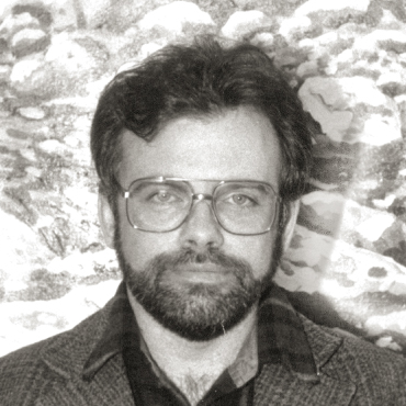 Black and white portrait of Michael Coyne with a beard and glasses, wearing a jacket over a collared shirt, facing the camera with a neutral expression.