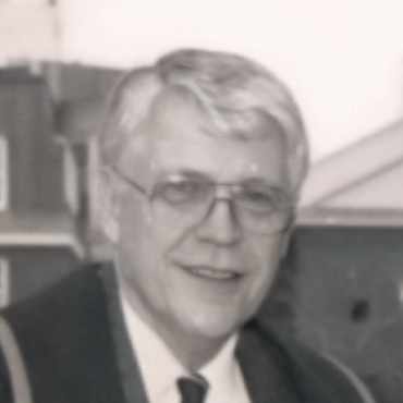 Black and white portrait of Noel Veitch with short gray hair and glasses, wearing a suit and tie, smiling slightly with a building blurred in the background.