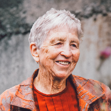 A colorful portrait of Penny Allderdice. She is wearing an orange-brown colored coat, with a red sweater underneath. She is smiling.