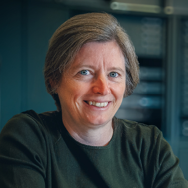 Portrait of Priscilla Renouf smiling, wearing a dark green top, photographed indoors with softly blurred shelves and equipment in the background.