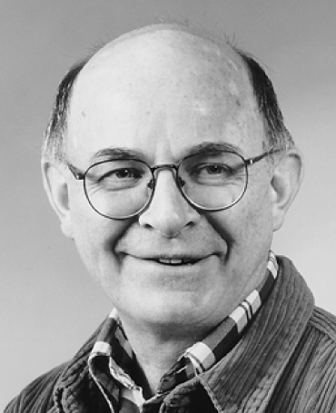 Black and white portrait of Neil Rosenberg, a man with round glasses, smiling slightly. He is wearing a plaid shirt under a corduroy jacket. The background is a plain, neutral tone.