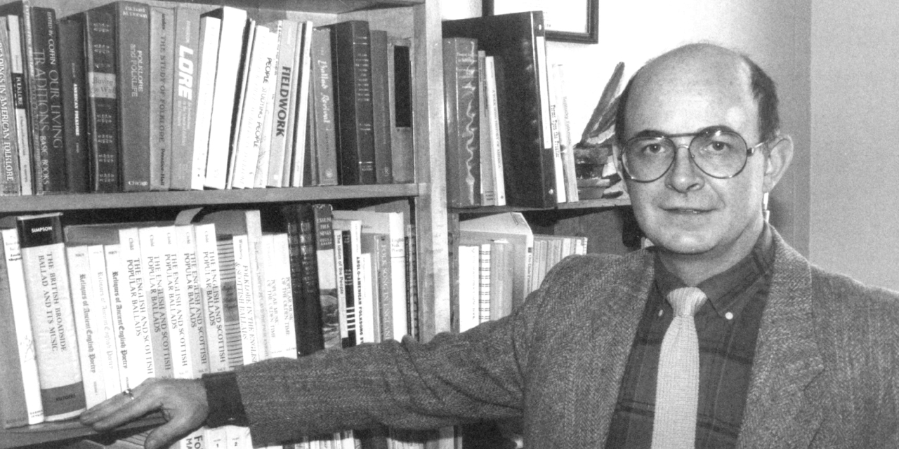 Black and white photograph of Neil Rosenberg, a bald man with round glasses, wearing a tweed jacket, plaid shirt, and a light-colored tie. He is standing with one hand resting on his office bookshelf, which is filled with academic books.