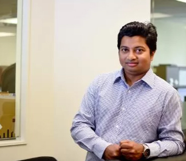 Kshama Roy, a man with short black hair and a slight smile, stands in an office setting, leaning slightly forward with his hands clasped. He wears a light-colored checkered button-up shirt. The background features office cubicles and a glass window with a partial reflection of the workspace. The lighting is bright and even.