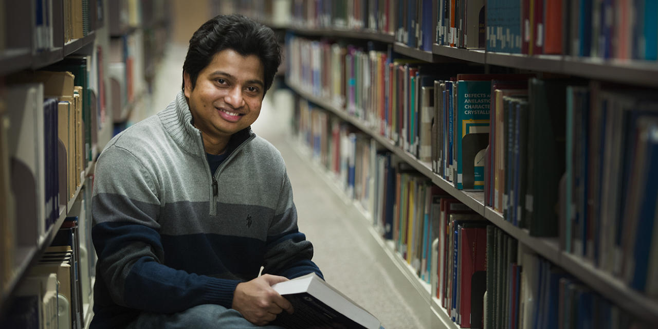 Kshama Roy, a man with short black hair and a warm smile, sits in a library aisle holding an open book in his hands. He wears a gray and navy-blue zip-up sweater. The shelves on either side of him are filled with books, creating a cozy and academic atmosphere. The lighting is soft, emphasizing the quiet and focused setting.