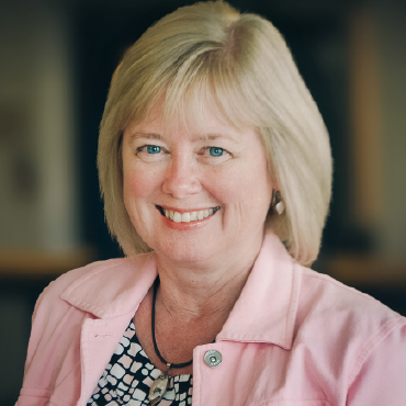 Head shot of Sonja Knutson wearing a pink jacket over a black, white and pink patterned blouse. She’s smiling happily.