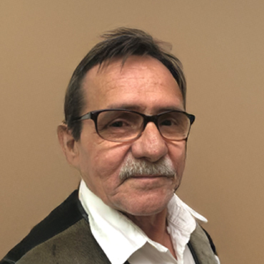 Color portrait of Timothy McNeill with short dark hair and mustache, wearing glasses, a white collared shirt, and a jacket, standing against a plain background.