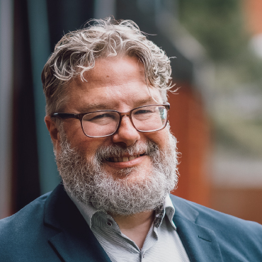 A colorful portrait of Tom Cooper. He is wearing a suit and is smiling. The background is blurred. The picture looks like it was taken during the daytime.