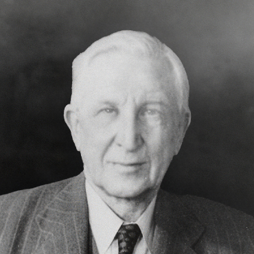 A black and white portrait of Vincent Burke. The background is blurred. He is wearing a suit and a tie and is smiling.