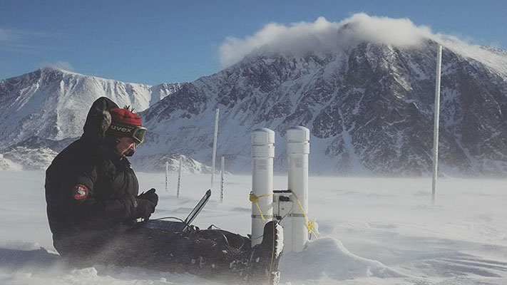 A man in dressed in winter clothing sitting on the snow with a laptop and research equipment. He is surrounded by blowing snow and mountains.