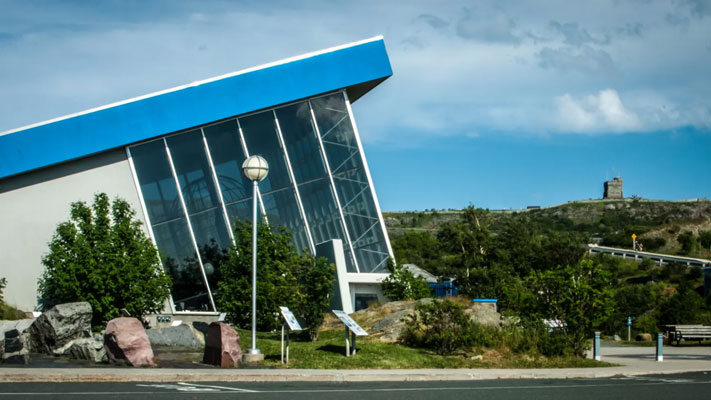 A photo of outside of the Johnson Geo Centre. The building is blue and white with mostly long vertical glass panes and is slanted.