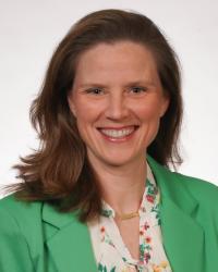 Ann Blackmore has shoulder length brown hair and wears a green blazer over a floral blouse. She is smiling at the camera.