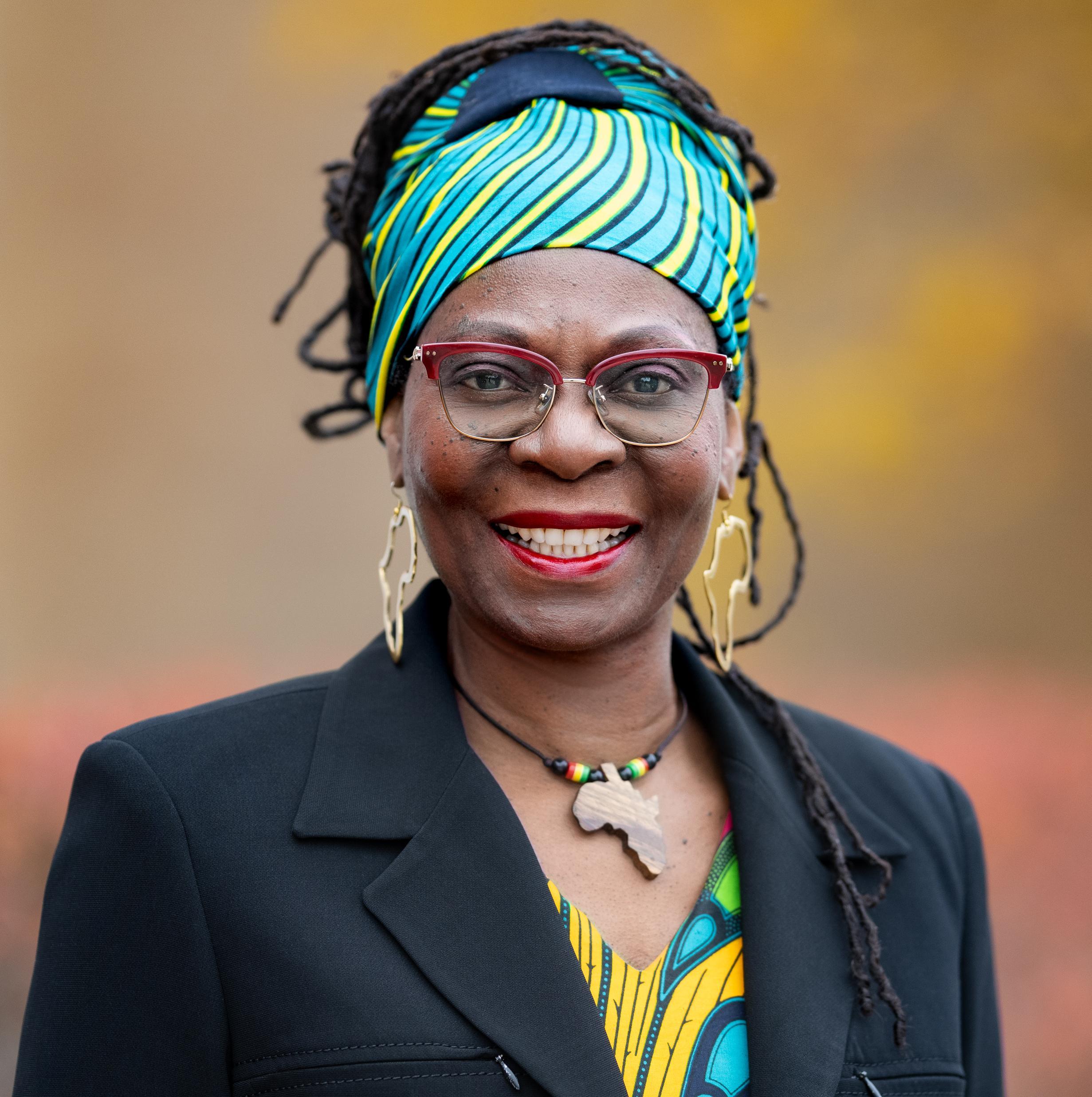 Dr. Delores V. Mullings, a Black woman wearing a green head wrap and a black blazer, looks at the camera and smiles.