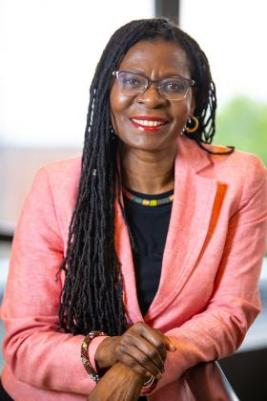 Dr. Delores V. Mullings, a Black woman wearing glasses and a pink blazer, is seen from the waist up. She is looking at the camera and smiling.