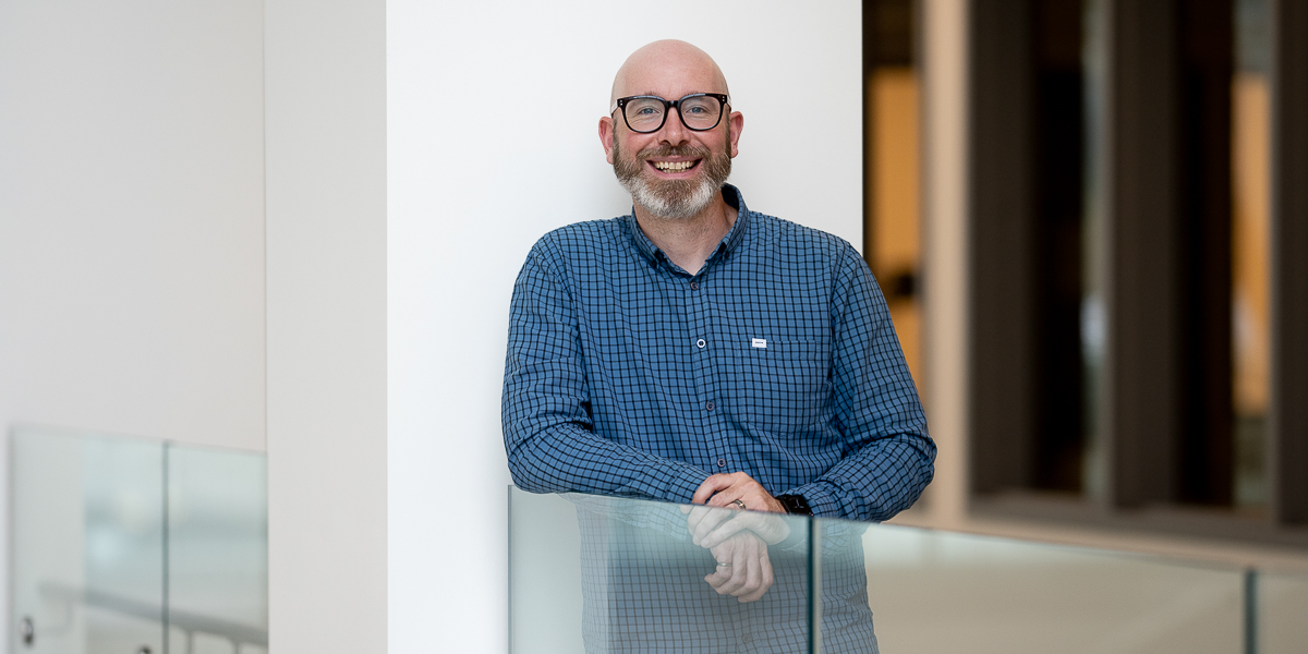 Man wearing glasses leaning against a glass railing.