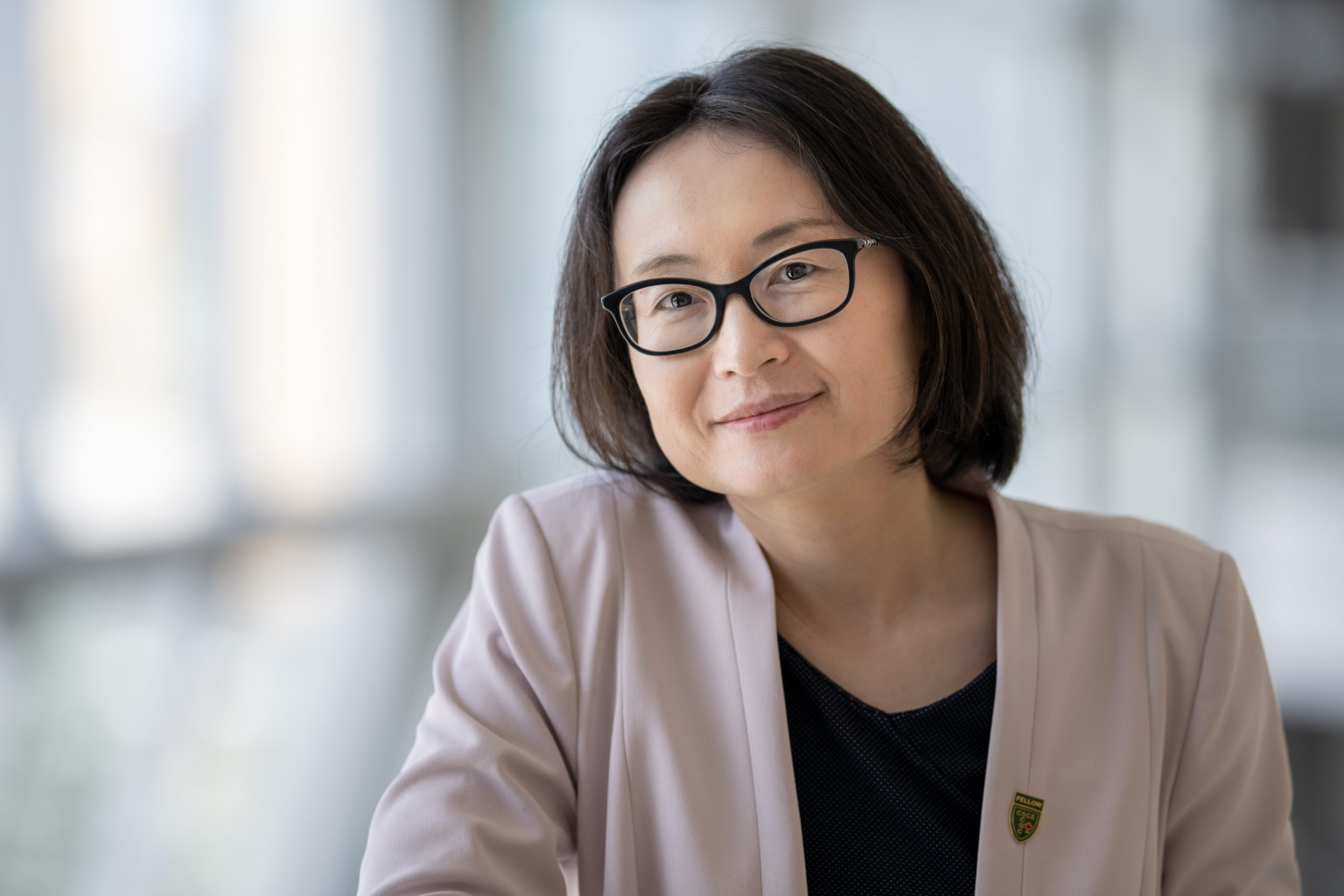 Woman with dark hair and glasses in pale pink blazer leans on railing.