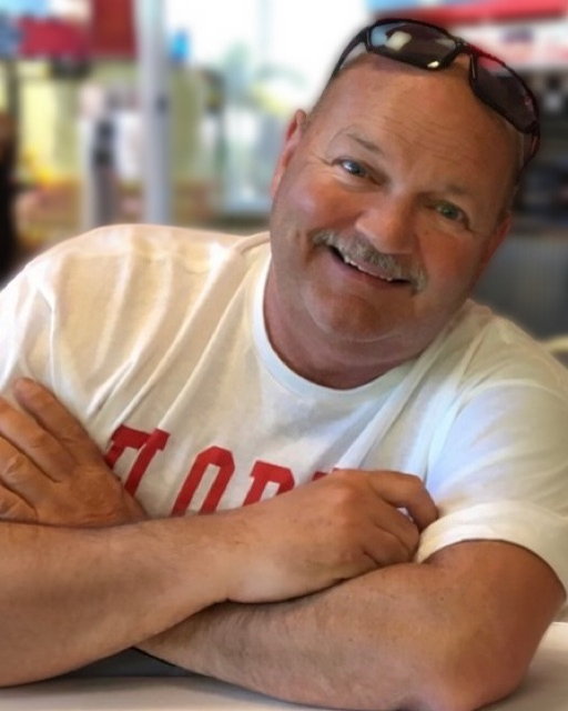 Middled-aged Caucasian man in white t-shirt, arms folded, with moustache, and smiling directly at the camera