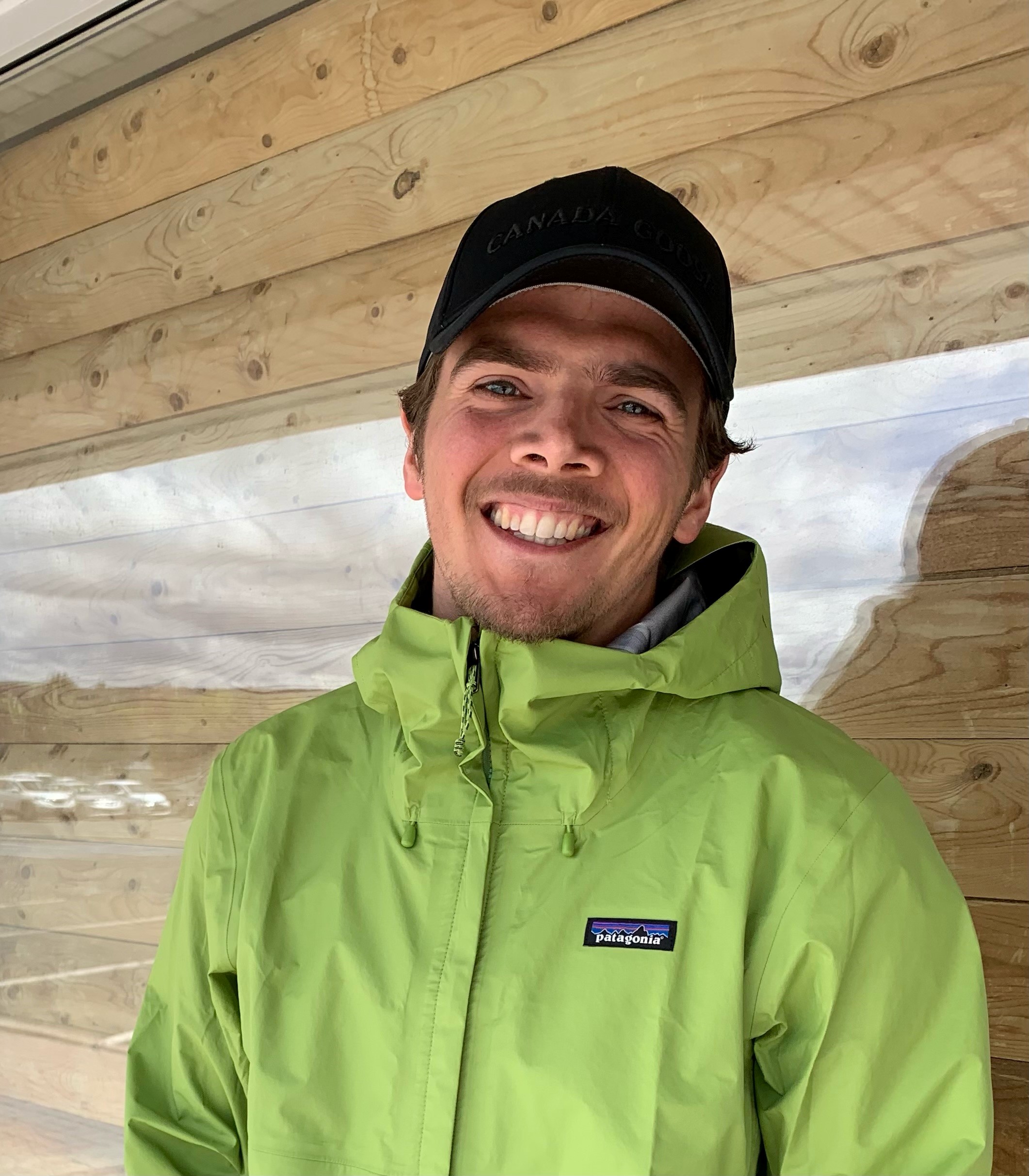 A young, Caucasian man with a big smile wearing a green jacket and a backwards ball cap.