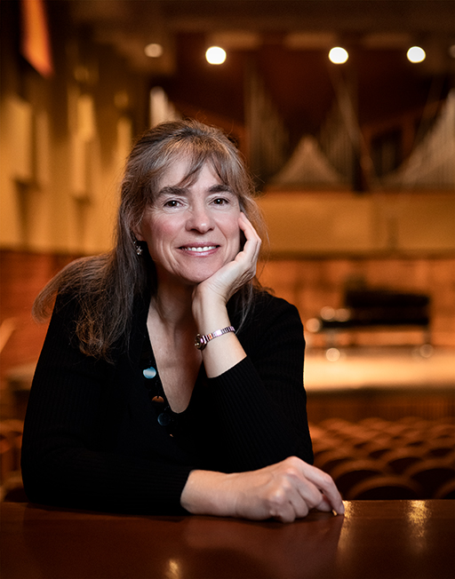 A woman who appears to be in her mid or late 40s, long blond hair, Caucasian complexion, smiling at the camera with chin resting in her hand. The background is the Cook Recital Hall, with a piano onstage in the distance.