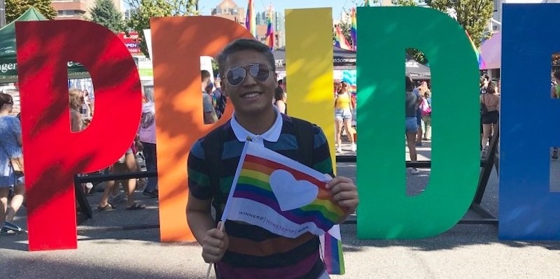 Nel Jayson Santos, who is smiling and holding a Pride flag, stands outside in front of large letters spelling 'pride'.