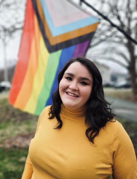 Brown haired woman in yellow shirt smiles in front of Pride flag