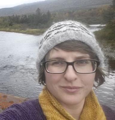 Picture of Joanne, a smiling woman wearing a knit hat, scarf, and sweater is outside by a river and trees