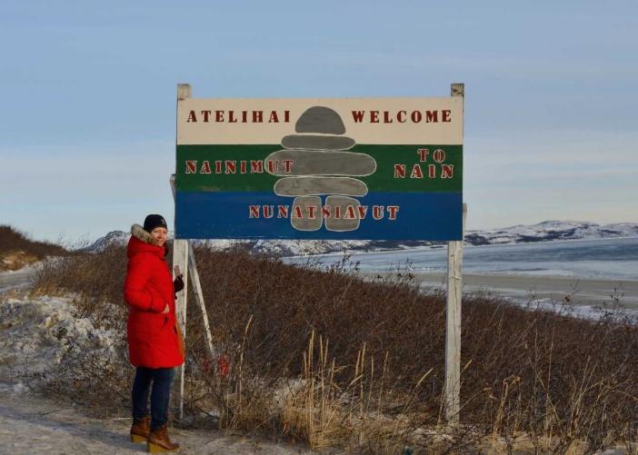 Valeri Pilgrim is standing beside a large sign that says Atelihai Nainimut, Nunatsiavut. Welcome to Nain, Nunatsiavut. The sign has a grey inukshuk in the middle on a white, green and blue background. The woman is outside, there is snow on the ground and the hills and the low plants are brown. 