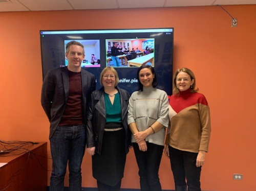 Dr. Nicolas Smith, myself, Dr. Lydia Goff and Dr. Laurie Twells stand next to each other in a row, smiling and looking at the camera. The wall behind them is orange and has a large television screen mounted on it.