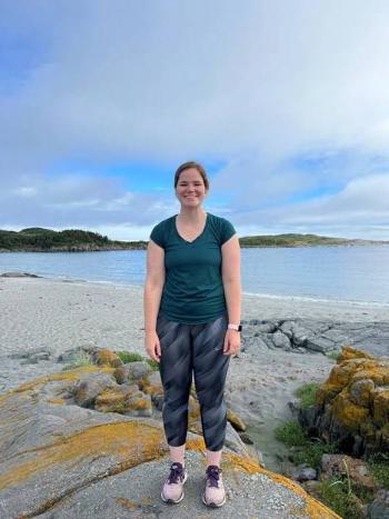 A woman, wearing a t-shirt and yoga pants, stands on a rocky shoreline with the ocean and a partly cloudy sky in the background.