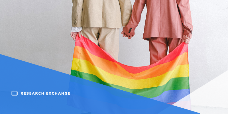 Photo of two people holding a pride flag. They are wearing beige and pink suits while holding hands.