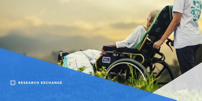 Picture of someone in a wheelchair on a grassy hill, their caregiver is tipping the chair back, and they are both watching the sunset.