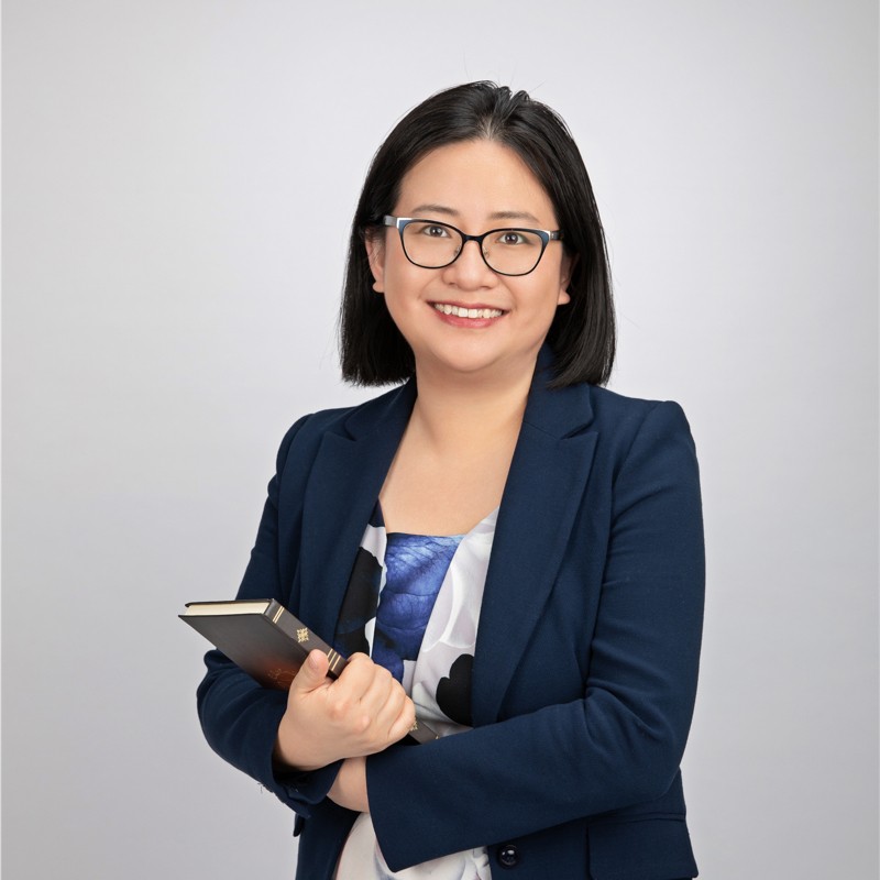 A woman with glasses and short dark hair in a blue blazer and white and blue blouse smiles as she holds a book